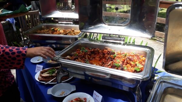 Un buffet généreux est dressé avec des plats chauds lors de la remise des prix LanKRéol 2017. Une main dépose des légumes sautés dans une assiette. Les plats sont présentés dans des bacs en inox sur une nappe bleue avec des étiquettes indiquant les noms des plats