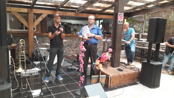 Un intervenant en tenue noire prend la parole au micro sur la scène décorée de fleurs lors de la remise des prix LanKRéol 2017, avec un trombone posé à ses côtés. Un autre intervenant en chemise bleue se tient debout à l'arrière-plan à côté des enceintes."