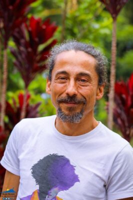 Un homme souriant, portant un t-shirt blanc, dans un jardin avec des plantes tropicales.
