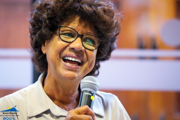 Une femme portant des lunettes sourit en tenant un micro.
