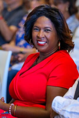 Une femme en robe rouge souriant assise parmi le public lors d'un événement.