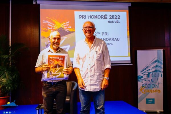 Deux hommes souriants sur scène, l'un tenant une affiche du Pri Honoré 2022