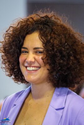 Une femme avec des cheveux bouclés sourit, vêtue d'une veste violette.