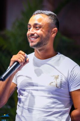 Un homme souriant tenant un micro, vêtu d'un t-shirt blanc.