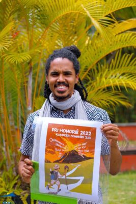Kaf marron souriant, tenant une affiche du Pri Honoré 2022.