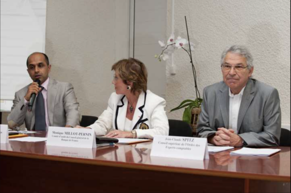 Trois intervenants assis à une table, dont un prend la parole avec un micro, lors d'une conférence.