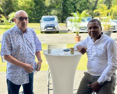 Jean françois Beaulieu et un membre de la déléguation de Mayotte debout près d'une table haute à l'extérieur, chacun tenant une tasse de café. L'un porte une chemise fleurie, l'autre une chemise blanche. Des voitures et une végétation luxuriante se trouvent en arrière-plan.