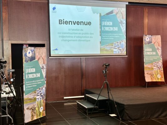 Une scène avec un écran projetant un message de bienvenue à l'atelier de co-construction sur les trajectoires d'adaptation au changement climatique. Deux panneaux sur les côtés affichent le thème 'La Réunion à l'horizon 2040'. Une caméra est installée devant la scène.
