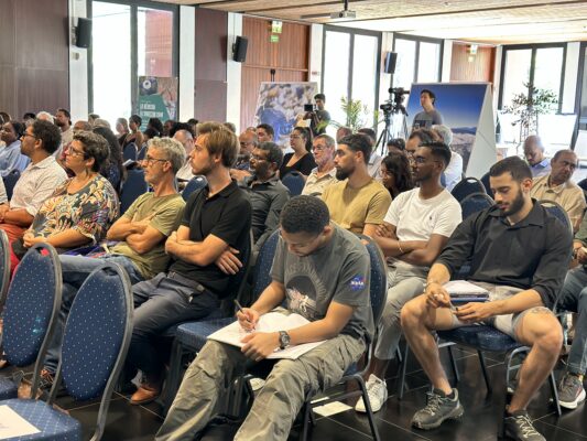 Un public diversifié assis lors d'une conférence, écoutant attentivement les interventions sur l'adaptation climatique. Certains participants prennent des notes, tandis que d'autres écoutent ou regardent les intervenants