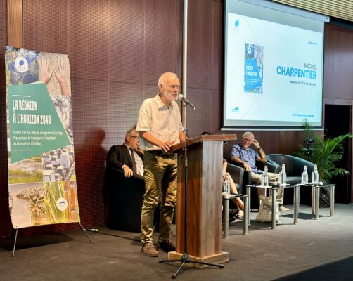 Michel Charpentier, membre du CCEE de Mayotte, parle lors d'une conférence avec un panneau sur l'adaptation au changement climatique intitulé 'La Réunion à l'horizon 2040'. Des membres du panel sont assis derrière lui.