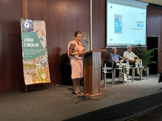 Huguette Bello, présidente de la Région Réunion, prend la parole lors d'une conférence sur 'La Réunion à l'horizon 2040'. Elle est debout derrière un pupitre, avec un écran projeté derrière elle affichant son nom. Un panneau à gauche présente les objectifs de l'événement liés aux défis climatiques. Deux hommes sont assis à côté d'elle sur la scène, écoutant attentivemen