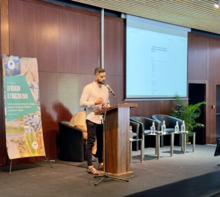 Valery GUERRIn en chemise blanche sur un podium, avec un écran derrière lui affichant une présentation. Un panneau à côté de lui mentionne 'La Réunion à l'horizon 2040', avec un message sur les défis climatiques.