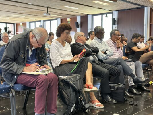 Participants d'une conférence, certains prennent des notes ou utilisent des ordinateurs portables. Un homme à l'avant-plan prend des notes dans un carnet, tandis que d'autres écoutent attentivement