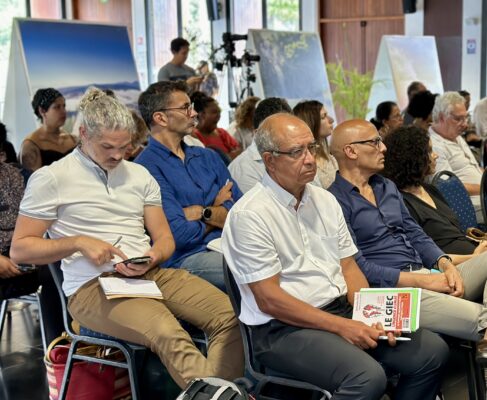 Un groupe de participants assis lors d'une conférence. L'un des hommes tient un livre intitulé 'Le GIEC : Une catastrophe annoncée'. D'autres personnes prennent des notes ou regardent leur téléphone.