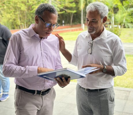 Félix lurel et un homme discutent tout en consultant un livre à l'extérieur. L'un porte une chemise rose à motifs et l'autre une chemise blanche, avec des lunettes accrochées à son col.