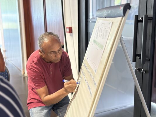 Un homme en t-shirt rouge écrit des notes sur un tableau papier lors d'un atelier collaboratif."