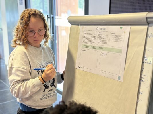 Une femme, tenant un stylo, se tient devant un tableau blanc sur lequel est affichée une feuille intitulée 'Biodiversité'. Elle semble en train d'écrire ou de participer à un atelier