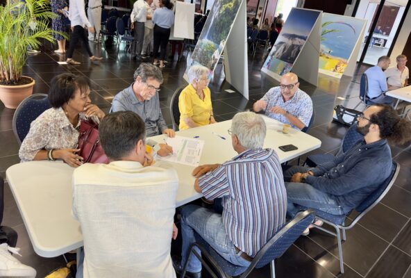 Un groupe de sept personnes est assis autour d'une table, discutant et prenant des notes lors d'un atelier collaboratif. En arrière-plan, des affiches présentant des paysages sont visibles
