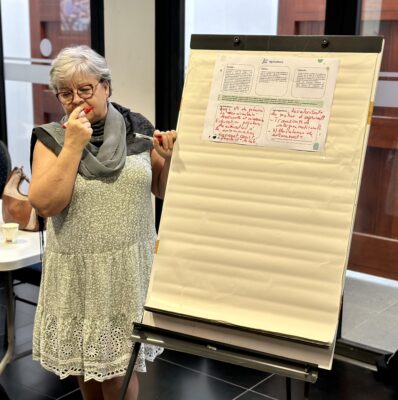 Une femme se tient à côté d'un tableau à feuilles mobiles lors d'un atelier. Elle réfléchit en tenant un stylo devant son visage. Sur le tableau, une feuille affiche des notes manuscrites en rouge sur un sujet lié à l'agriculture. Un sac à main et un gobelet sont visibles sur une table à l'arrière-plan.