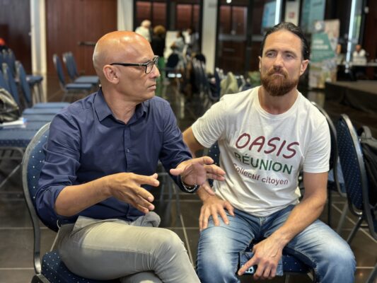Deux hommes discutent lors d'une conférence. L'un porte une chemise bleue, l'autre un t-shirt avec l'inscription 'Oasis Réunion - Collectif citoyen