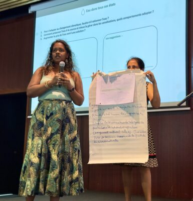 Deux femmes présentent les conclusions d'un atelier sur la gestion de l'eau. L'une tient un micro et l'autre montre un tableau papier avec des notes, avec une diapositive projetée en arrière-plan sur la question de l'adaptation de l'eau au changement climatique.
