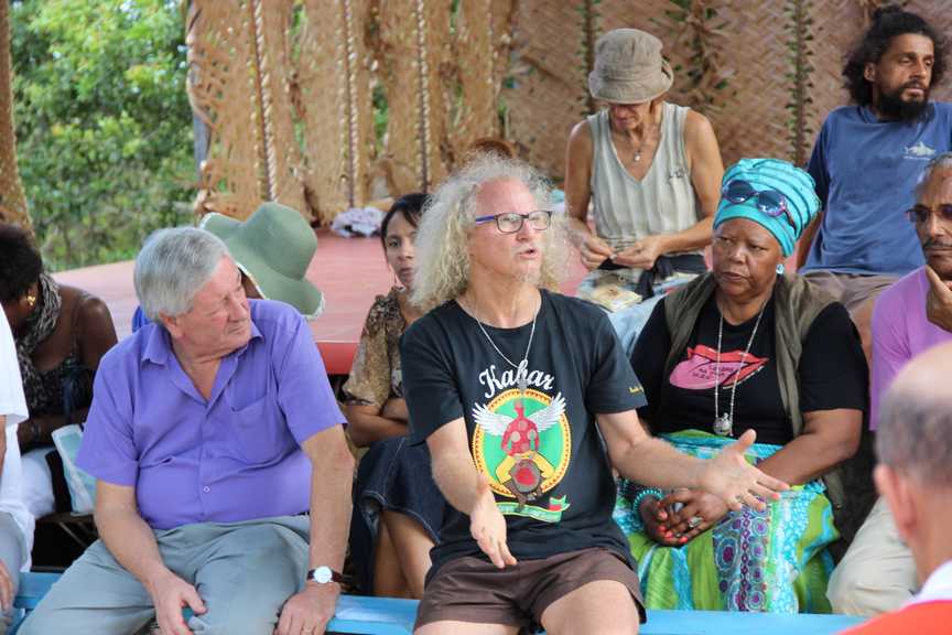 Daniel Waro, homme aux cheveux blancs, échange lors d’une rencontre conviviale en extérieur, entouré de plusieurs participants assis.