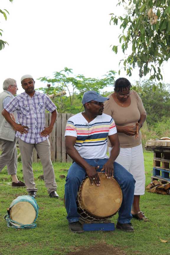 jean-pierre_bacot_et_rosemonde_de_neef_du_ccee_de_guyane jouant du tambour en extérieur, accompagné de deux participants discutant à l’arrière-plan.