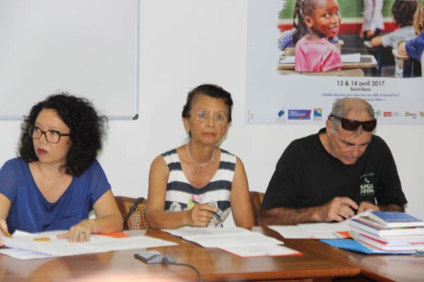 Trois membres du CCEE assis à une table, prenant des notes lors d’une réunion, avec des documents et des livres devant eux. Un poster éducatif est visible en arrière-plan.