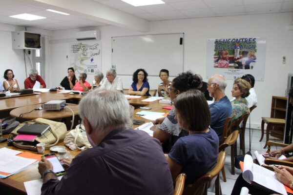 Vue d’ensemble d’une réunion du CCEE, avec des participants assis autour d’une grande table, échangeant et prenant des notes. Des affiches éducatives et un tableau blanc sont visibles en arrière-plan.