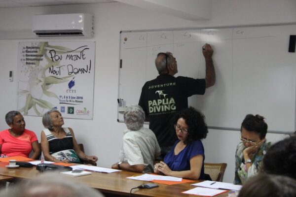 Un membre du CCEE écrit sur un tableau blanc lors d’une réunion, tandis que d’autres participants l’écoutent et prennent des notes autour de la table.