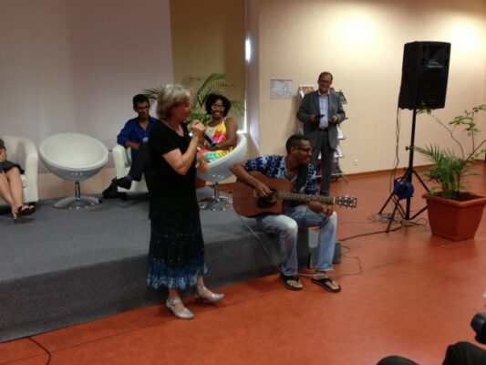 "Une intervenante en robe noire prend la parole au micro pendant la remise des prix LanKRéol 2013, accompagnée d'un guitariste assis à ses côtés sur scène. À l'arrière-plan, d'autres participants suivent la scène avec attention."