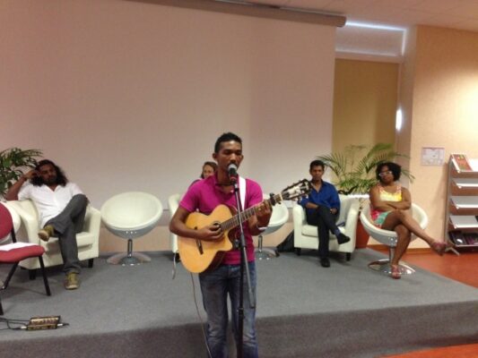 Un chanteur en chemise rose joue de la guitare et chante au micro sur scène lors de la remise des prix LanKRéol 2013. En arrière-plan, plusieurs intervenants assis suivent la prestation avec intérêt.