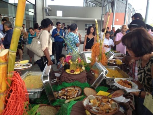 Un buffet coloré et garni de spécialités locales est dressé pour les participants lors de la remise des prix LanKRéol 2013. Des invités se servent autour des plats disposés sur une table décorée avec des cannes à sucre et des fruits tropicaux, tandis que d'autres discutent à l'arrière-plan."
