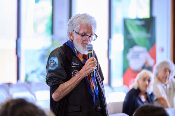 Homme intervenant avec un micro lors d'une conférence avec des participants en arrière-plan.