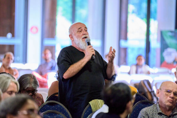 Homme intervenant avec un micro lors d'une conférence, portant un tee shirt noir avec des participants en arrière-plan.