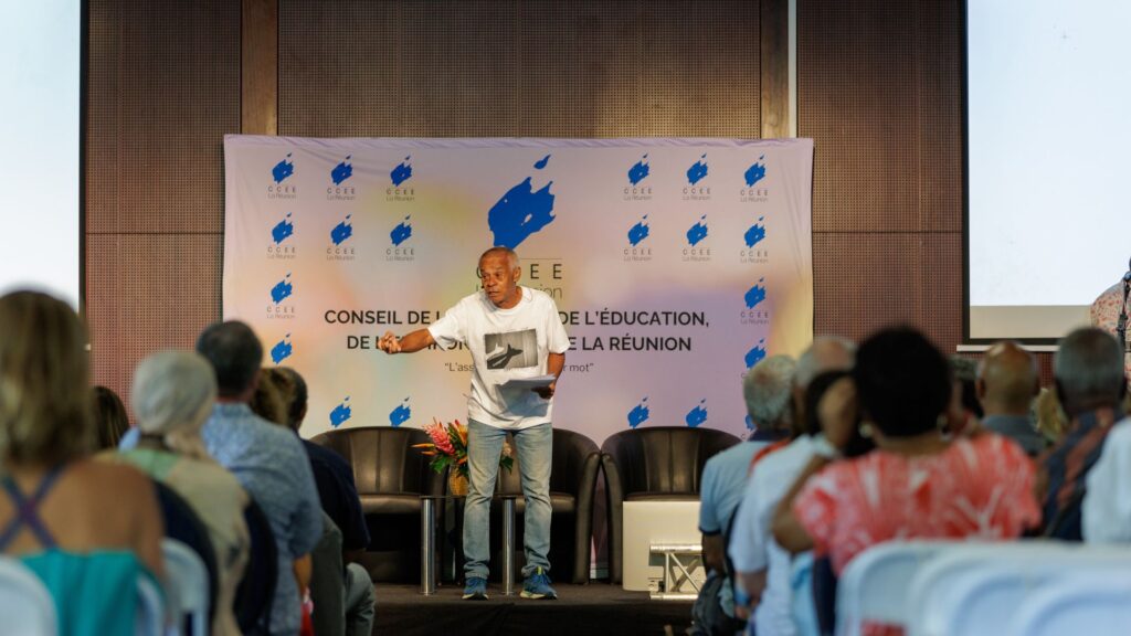 Sully Andoche, vêtu d'un t-shirt blanc avec un imprimé et un jean, debout sur scène devant une banderole du CCEE, en train de réciter un texte face à un public attentif lors des 40 ans du CCEE