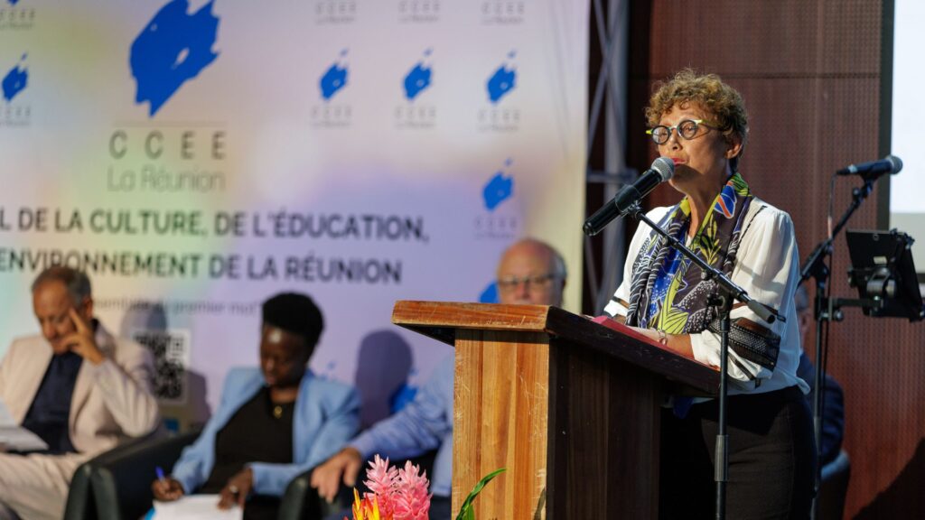 Marie-Jo Lo Thong, directrice de la DAC, habillée d'une blouse blanche avec un foulard coloré, parlant au micro derrière un pupitre en bois, lors des 40 ans du CCEE. En arrière-plan, des officiels assis, prenant des notes