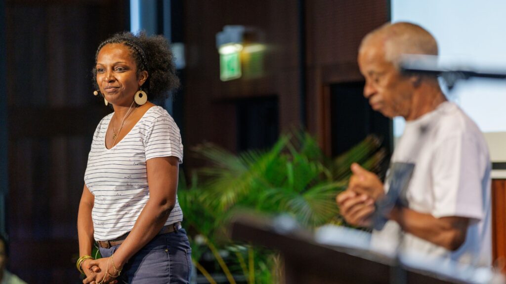 Alexandrine Dijoux, vêtue d'un t-shirt rayé blanc et d'un pantalon bleu, s'exprimant sur scène avec un micro-cravate lors des 40 ans du CCEE. Sully Andoche est visible flou à l'arrière-plan.