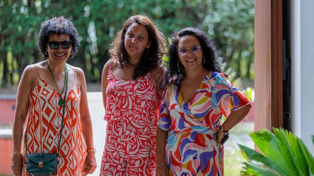 Photo de trois femmes souriantes, habillées en robes à motifs colorés, posant à l'entrée d'une salle lors de l'événement des 40 ans du CCEE, avec un arrière-plan de nature et de lumière.