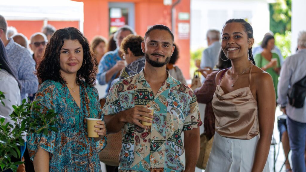 Photo de trois jeunes adultes souriants, vêtus de tenues élégantes et colorées, lors de l'événement des 40 ans du CCEE. En arrière-plan, des participants discutent autour de boissons dans un cadre lumineux.