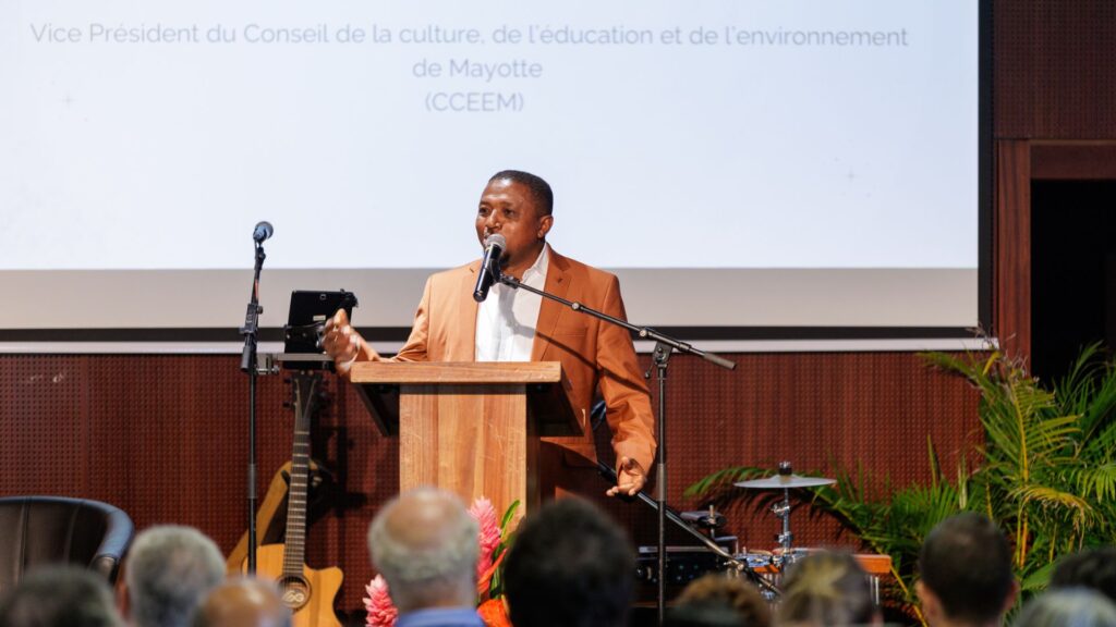 Ali Madi, vêtu d'une veste marron clair et d'une chemise blanche, prenant la parole au micro sur une estrade en bois, avec un écran projetant son nom et sa fonction lors des 40 ans du CCEE.