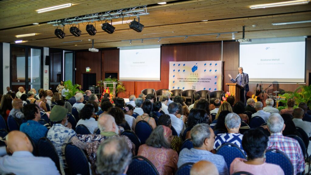 Photo large de la conférence des 40 ans du CCEE avec un public nombreux assis, écoutant le discours de Rostane Mehdi sur scène, dans une salle ornée de panneaux en bois et équipée d'un écran de projection