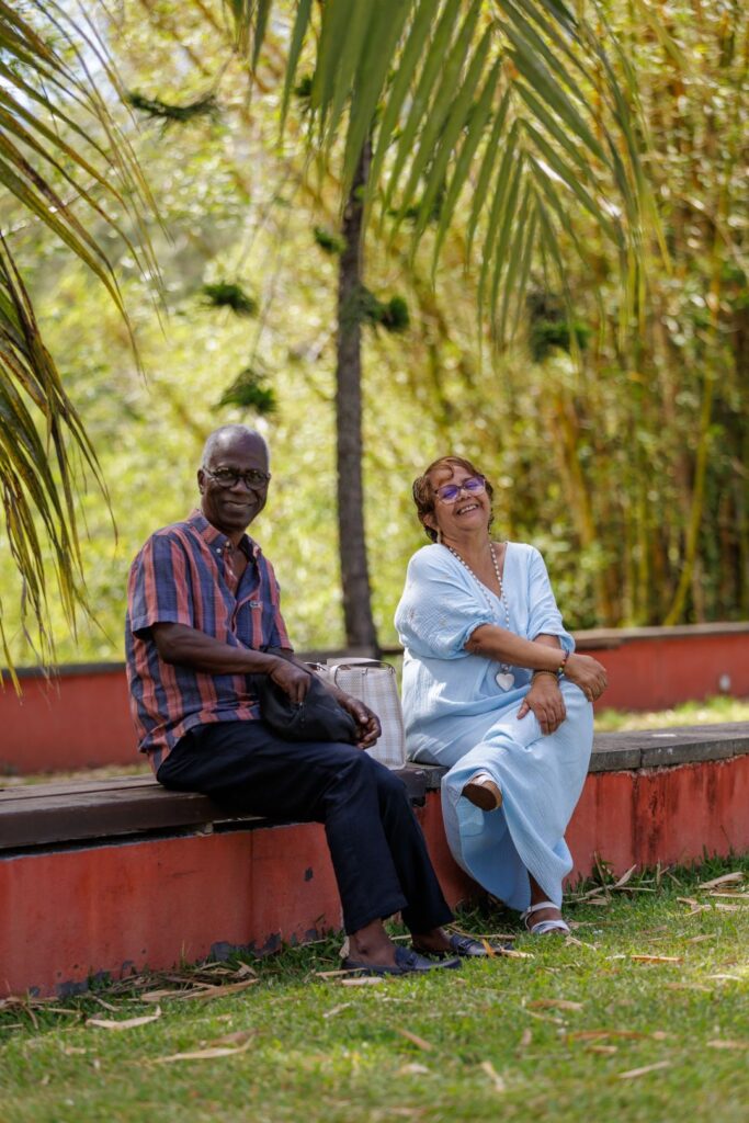 Un homme en chemise rayée rouge et bleue assis à côté d'une femme en robe bleue claire, souriant sur un banc en pierre, entourés de palmiers et de bambous, lors des 40 ans du CCEE.