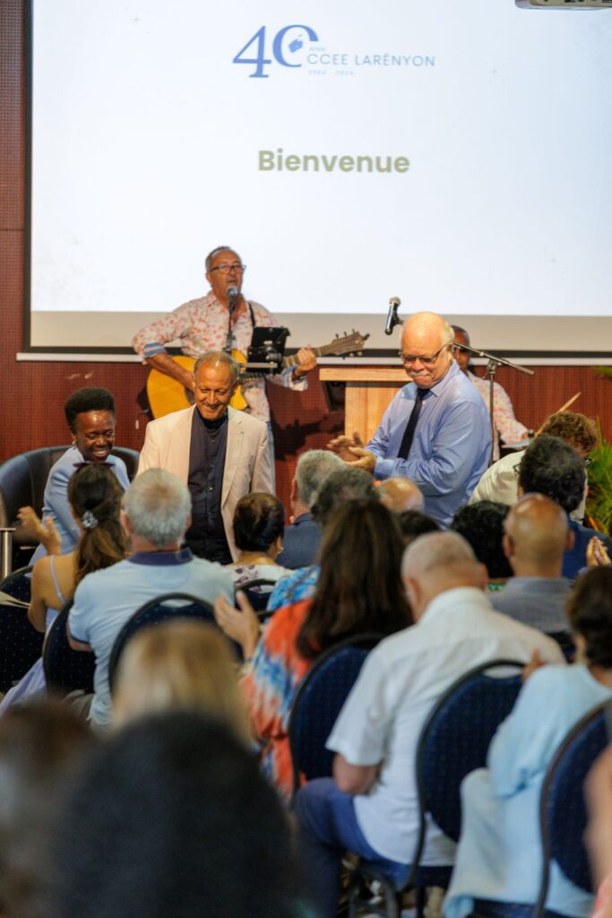 Devant la scène, Patricia Profil en costume bleu, Roger Ramchetty en veste claire et David André applaudissant lors de la cérémonie d'ouverture des 40 ans du CCEE, avec un musicien jouant de la guitare en arrière-plan.