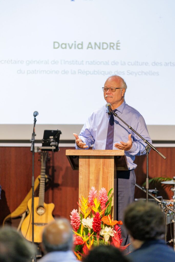 David André, vêtu d'une chemise bleu clair et d'une cravate, s’exprimant au micro depuis un pupitre en bois, avec des fleurs tropicales en décoration et un écran projetant son nom et sa fonction, lors des 40 ans du CCEE