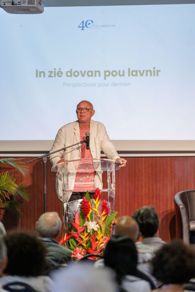 Dominique Carrère, vêtu d'une veste beige et d'un t-shirt rose, s’exprimant au micro derrière un pupitre transparent avec des fleurs tropicales, lors de la présentation 'In zié dovan pou lavenir' pendant les 40 ans du CCEE.