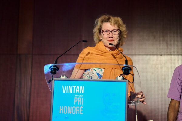 Une femme portant une écharpe orange prend la parole derrière un pupitre affichant le visuel Vintan Pri Honoré 2024. Elle s'adresse à l'audience avec un sourire chaleureux.