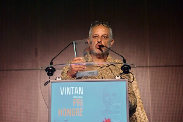 Un homme, vêtu d'une chemise à motifs, est debout sur scène et tient un trophée Vintan Pri Honoré 2024. Il est positionné derrière un pupitre portant l'affiche de l'événement.
