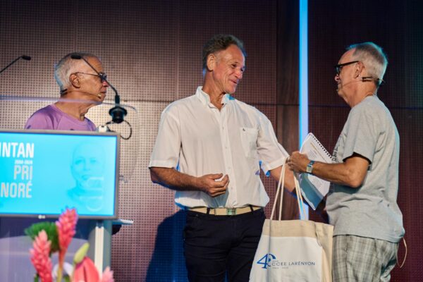 "Lors des Vintan du Prix Honoré, trois intervenants échangent sur scène. À gauche, Dominique Carrère en t-shirt rose est attentif. Au centre, un homme en chemise blanche sourit tout en tendant la main. À droite, un autre intervenant en t-shirt gris, tenant des notes, lui remet un sac commémoratif du CCEE La Réunion.