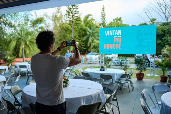 Un participant en t-shirt blanc prend une photo avec son téléphone portable lors des Vintan du Prix Honoré. Devant lui, des tables rondes dressées avec des nappes blanches et des chaises sont disposées en extérieur. À l'arrière-plan, un grand écran affiche le texte 'Vintan 2000-2020 Pri Honoré' sur un fond bleu avec un portrait en filigrane. La scène est entourée de verdure et de plantes.